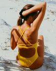 JAYD model Lauren sitting on a beach in the Ignite yellow Airlie one piece swimsuit with a semi adjustable cross-back straps for maximum comfort and support.