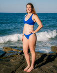 A woman wearing a bright blue two-piece bikini stands on dark rocks at the ocean's edge, with waves and blue sky in the background.