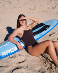 JAYD Swimwear model Cyra wearing the Apsley Cacao swimsuit with white shoulder straps, reclines on a blue and white ski on a sandy beach, shielding her eyes from the sun.