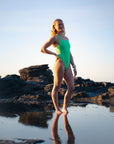 JAYD athlete Emma wearing the bright green Meadow Apsley one-piece swimsuit stands barefoot in shallow water on a rocky beach at sunset, with her reflection visible on the wet surface.