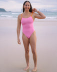 JAYD Swimwear athlete Jenaya Massie standing on a beach wearing the Airlie one piece swimsuit in Fairy Floss pink with contrasting Flare orange shoulder straps.