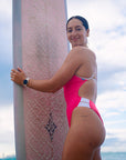 Athlete Jenaya Massie standing on the beach holding her surf board, ready to hit the waves in her JAYD one piece Ibiza swimsuit in vibrant pink sorbet with contrasting white hip cut-outs and back straps.
