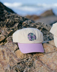 Cream and purple JAYD Swimwear Baseball Cap sitting on a rock at the beach.
