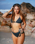 JAYD model Cass walking on a beach listening to a vintage music player, wearing the Kiama bikini top in Carbon grey with matching Carbon bottoms.