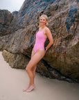 JAYD Swimwear model on a sandy beach leaning against a large rock wall, wearing the bright pink fairy floss Tallow one-piece swimsuit.