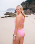 JAYD model stands on a sandy beach wearing a bright pink fairy floss fixed-back, full bum coverage Tallow one-piece swimsuit, looking back over her shoulder. Rocky cliffs and the ocean are visible in the background under an overcast sky.
