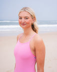 JAYD model stands on a sandy beach wearing the bright fairy floss pink Tallow one-piece swimsuit. The ocean and pale sky are visible in the background.