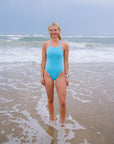 JAYD model standing on a sandy beach with ocean waves behind her wearing the Tallow one piece swimsuit in Mykonos blue.