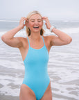 JAYD Swimwear model laughing on a beach standing in shallow ocean water, wearing the Tallow one piece swimsuit in Mykonos blue.