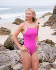 JAYD Swimwear model standing on a rocky beach with the ocean in the background, wearing the Tallow one piece swimsuit in orchid pink.