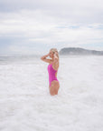 JAYD Swimwear model stands in foamy ocean waves wearing the bright pink orchid Tallow one-piece swimsuit, with a cloudy sky and distant shoreline in the background.