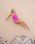 JAYD model wearing the orchid one-piece Tallow swimsuit sits on a sandy beach, viewed from above. Rocks are visible in the foreground.