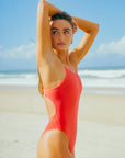 Triathlete Lauren wearing the Blaze red Zimmy one piece swimsuit standing on a beach with ocean and sky in the background