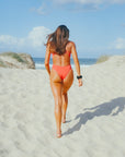 JAYD model walking down the beach towards the water in the fiery red Blaze one piece Zimmy swimsuit.