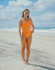Triathlete Charlotte standing on a beach wearing the JAYD Swimwear Zimmy one piece in Flare orange.