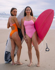 JAYD Swimwear athletes Lauren and Jenaya standing on a beach holding surfboards, wearing the inverted matching Airlie one piece swimsuits in flare orange and fairy floss pink.