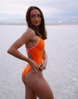 JAYD model Lauren wearing the Airlie one piece swimsuit in flare orange with contrasting fairy floss pink shoulder straps, standing on a beach looking over her shoulder.