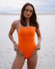 Triathlete Lauren Kirwick standing on a beach wearing the JAYD Swimwear Airlie one piece in flare orange with contrasting fairy floss pink shoulder straps.