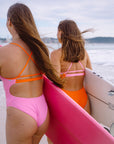 Two JAYD Swimwear athletes on a beach seen from behind, wearing bright fairy floss pink and flare orange Airlie one-piece swimsuits and carrying surfboards.