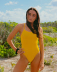 Athlete Lauren posing in the JAYD Swimwear Airlie one piece swimsuit in Ignite yellow on a beach with green shrubbery behind her.