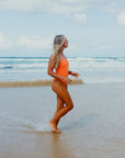 Woman in the JAYD Swimwear Albi one piece Flare orange swimsuit walking on a beach with ocean waves in the background.
