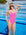 JAYD Swimwear model standing in front of a pool wearing the pink sorbet Albi one piece swimsuit with contrasting Mykonos blue shoulder straps.