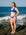 A woman wearing a bright blue two-piece bikini stands on dark rocks at the ocean's edge, with waves and blue sky in the background.