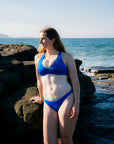 A woman wearing a bright blue two-piece bikini stands on dark rocks by the ocean, with waves and a pale blue sky in the background.