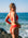 JAYD Swimwear model Jemima wearing the Amalfi bikini bottoms in ruby red, stands on a rocky beach with the ocean and blue sky behind her, looking over her shoulder.