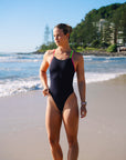 JAYD Swimwear athlete Claudia Slaven wearing the black Apsley one-piece swimsuit with bright pink sorbet and rave green shoulder straps walks on a sandy beach with ocean waves and trees in the background.