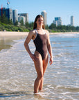 JAYD Swimwear athlete Cyra wearing the dark brown cacao Apsley one-piece swimsuit with contrasting white shoulder straps, standing in shallow ocean water on a beach, with sand, greenery, and high-rise buildings visible in the background under a clear blue sky.