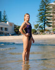 Surf Life Saving athlete Brodee Trinca standing in shallow ocean water on a beach wearing the JAYD Swimwear Apsley one piece swimsuit in Acai with contrasting fairy floss and sangria shoulder straps.