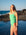Surf life saving athlete Emma Woods wearing the Meadow green JAYD Swimwear Apsley one-piece swimsuit stands on a rocky beach with the ocean and blue sky in the background.