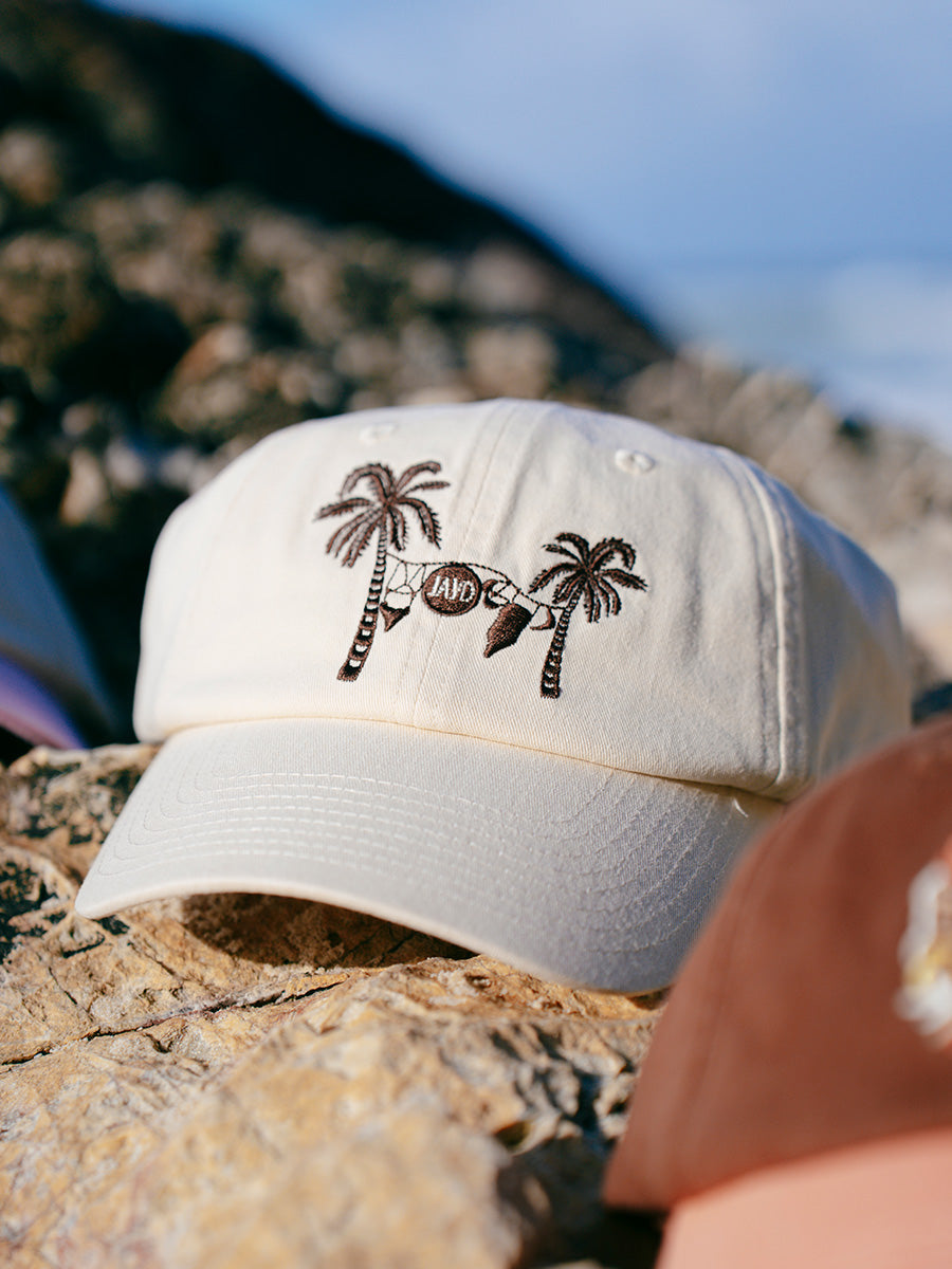 Cream JAYD Swimwear baseball hat with brown palm trees &amp; swimsuit embroidery sitting on a rock.