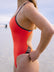 JAYD model wearing the bold Lennox one-piece swimsuit in blaze with contrasting brown cacao shoulder straps and hip panels, standing outdoors near the water with a blurred beach in the distance.