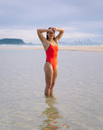 JAYD Swimwear model Kate wearing the Blaze Lennox one-piece swimsuit with contrasting cacao shoulder straps and hip cut-outs standing in shallow water at a beach, with her hands on her head, and a distant city skyline and island visible in the background.
