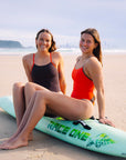 JAYD Swimwear athletes Molly and Kate sitting on a green surf craft at the beach, wearing matching contrasting swimsuits in blaze and cacao.
