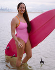 Ski paddler Jenaya Massie wearing the JAYD Swimwear Airlie swimsuit in fairy floss pink with contrasting flare orange shoulder straps, holding her pink surfboard on the beach.