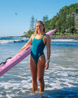 JAYD Swimwear model Brodee standing in shallow ocean water on a beach, wearing the Cove Finley one piece swimsuit with contrasting moss green shoulder straps, holding a pink ski.