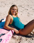 JAYD Swimwear athlete Brodee Trinca wearing the Cove Finley one-piece swimsuit with Moss green shoulder straps reclines on a pink ski on a sandy beach.