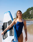 Surf life saving athlete Cyra wearing the navy one piece swimsuit with contrasting white shoulder straps, standing on a beach with her blue ski.