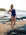 JAYD athlete Emma Woods wearing the navy blue Finley one-piece swimsuit stands on a rocky beach at sunset, with ocean waves and distant buildings in the background.