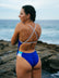JAYD model Madi standing on a rock at the beach wearing the Ibiza one piece swimsuit in cobalt blue with contrasting white semi-adjustable cross over back straps for maximum support & security, along with a full bum coverage.