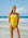 JAYD model standing on the beach in Ignite yellow Ibiza swimsuit with contrasting flare orange shoulder straps, standing on a beach with blue sky and ocean behind her.