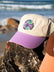 Salty Summer embroidery design on the cream JAYD Swimwear baseball cap with purple brim, sitting on a rock with the ocean in the distance.