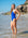 JAYD model wearing the bright blue cobalt one-piece swimsuit stands in shallow ocean water on a beach, with rocks and blue sky in the background.