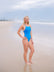 JAYD Swimwear model in a bright Cancun blue one-piece Tallow swimsuit on a sandy beach with the ocean and cloudy sky in the background.