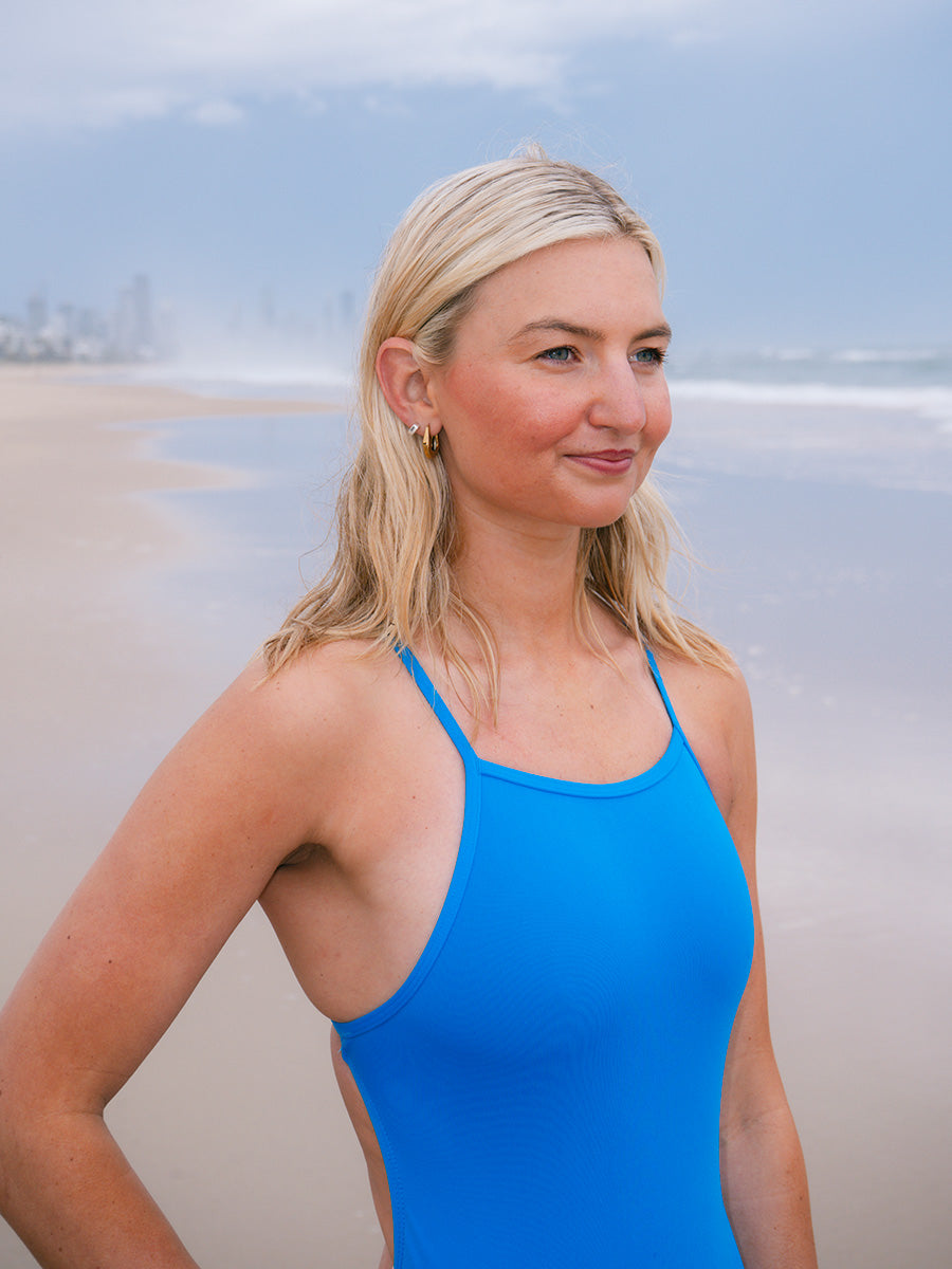 JAYD Swimwear model wearing a bright blue Cancun Tallow one-piece swimsuit stands on a sandy beach with the ocean and distant city skyline in the background.