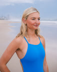 JAYD Swimwear model wearing a bright blue Cancun Tallow one-piece swimsuit stands on a sandy beach with the ocean and distant city skyline in the background.