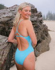 JAYD Swimwear model standing on a sandy beach next to large rocks and looking back over her shoulder, wearing the Tallow one piece swimsuit in Mykonos blue with triangle fixed-back straps and full bum coverage.